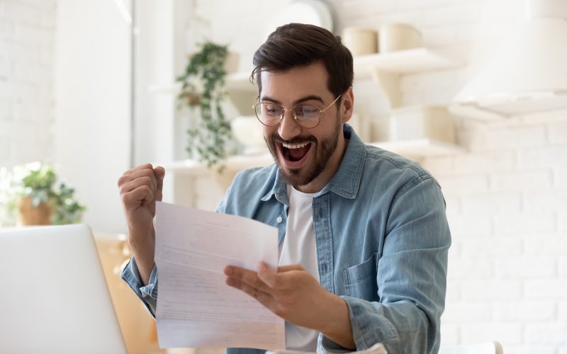Excited euphoric happy young man holding reading paper postal mail letter amazed overjoyed by good news, got new job celebrate taxes refund receive salary payment loan approval sit at home table